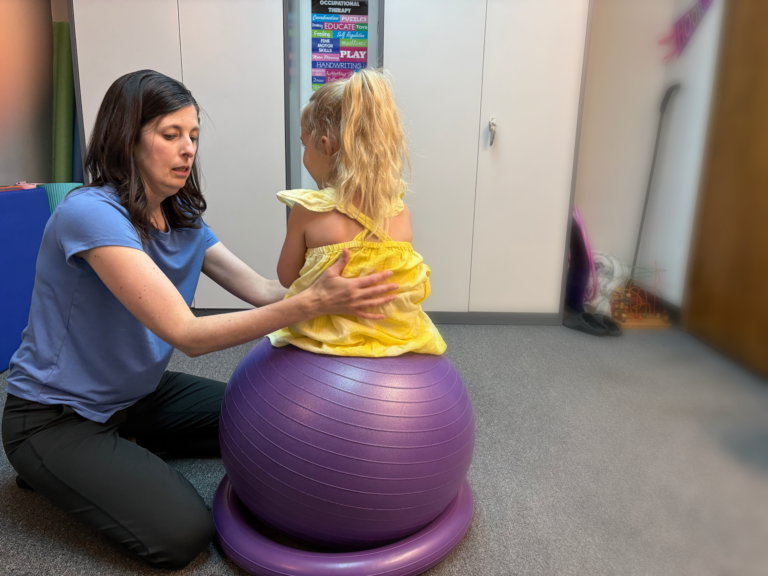A blond haired little girl in a yellow dress participates in occupational therapy while sitting on a purple balance ball