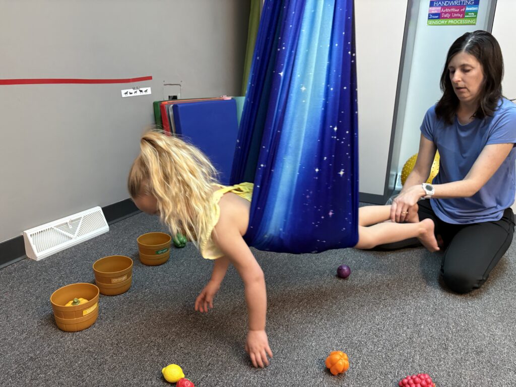 A blond haired little girl participates in motor occupational therapy sorting fruits while lying prone suspended by a body sling
